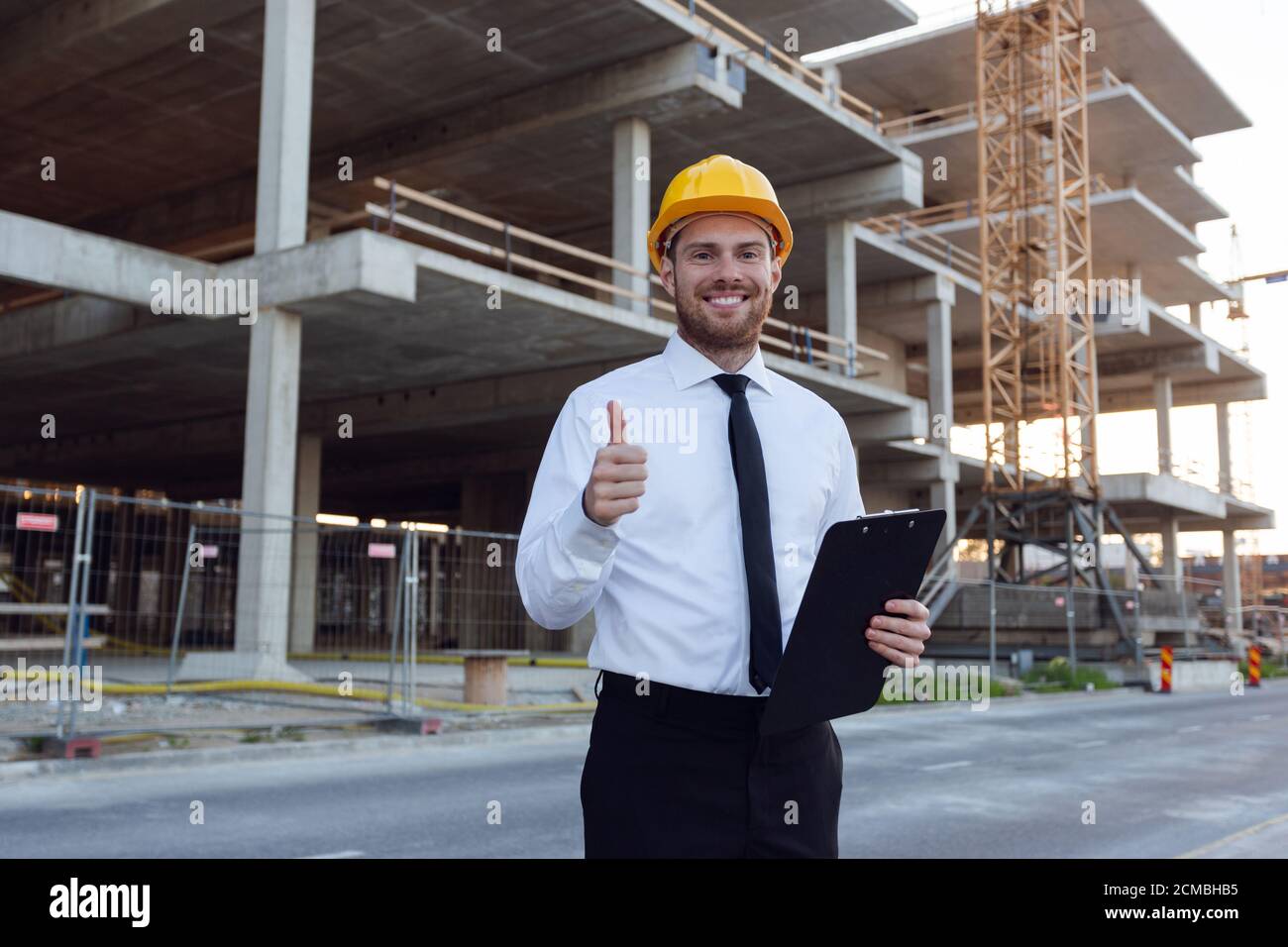 Geschäftsmann trägt HardHat Holding Zwischenablage in den Händen zeigt Daumen nach oben. Baumeister Controlling Haus Gebäude. Mann, der in der Nähe von Bauarbeiten schwelte Stockfoto