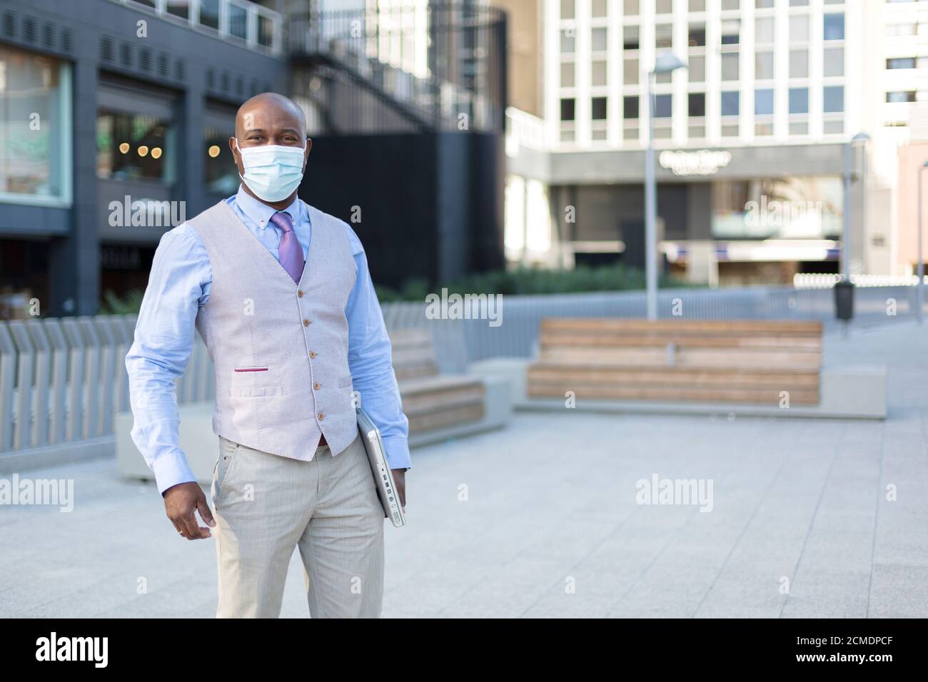Unternehmungslustiger schwarzer Mann auf der Straße. Er trägt eine Gesichtsmaske und hält einen Laptop unter seinen Arm. Konzept der neuen Normalität im Kontext der Coronavirus-Pandemie. Stockfoto