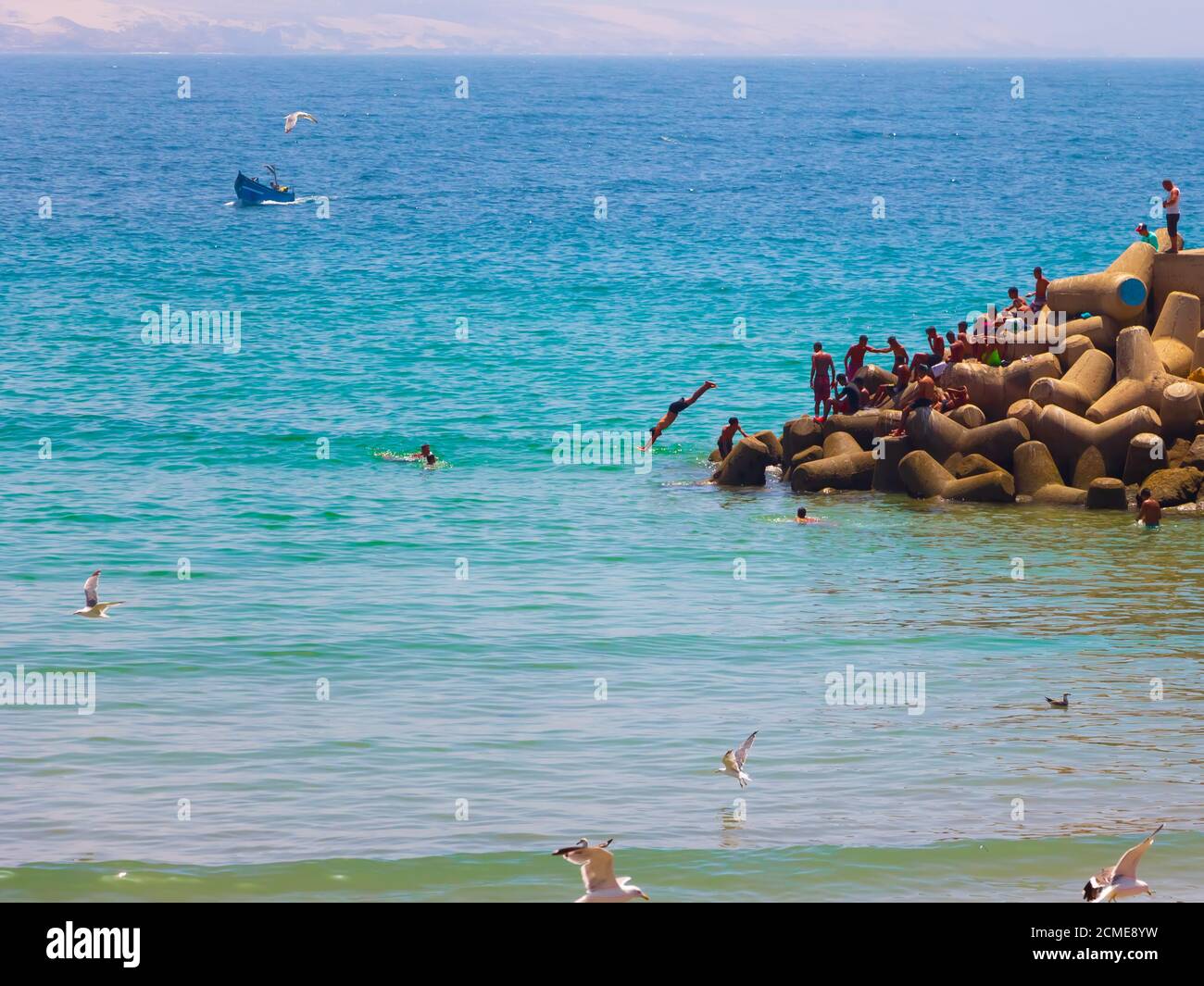 Agadir, Marokko - 14. AUGUST 2020 : Menschen schwimmen am Rande eines Hafens, im blauen atlantik, mit Möwen oben und einem blauen Boot Stockfoto