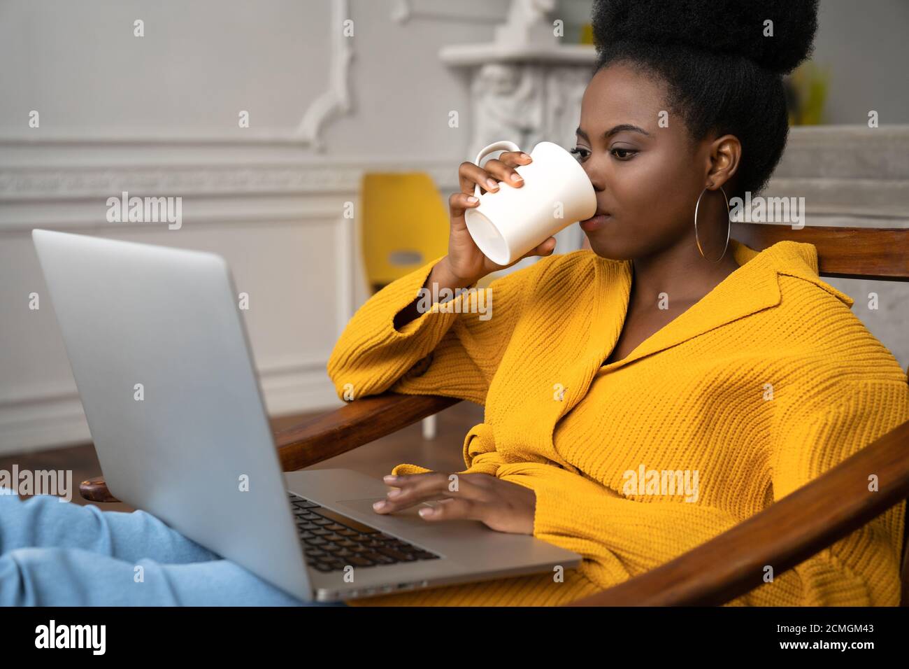 Afroamerikanische Biracial Frau mit Afro-Frisur in gelber Strickjacke sitzen auf Sessel, Fernstudium, Online-Arbeit auf Laptop, Videos ansehen Stockfoto