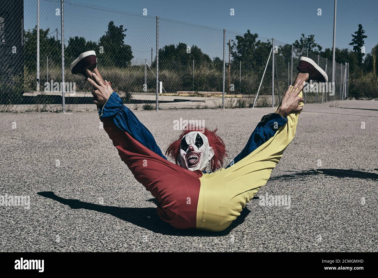 Ein gruseliger Clown, der ein gelbes, rotes und blaues Kostüm trägt, auf dem Asphalt auf seinem Rücken liegt, mit erhobenen Beinen und herausgestreckter Zunge draußen Stockfoto