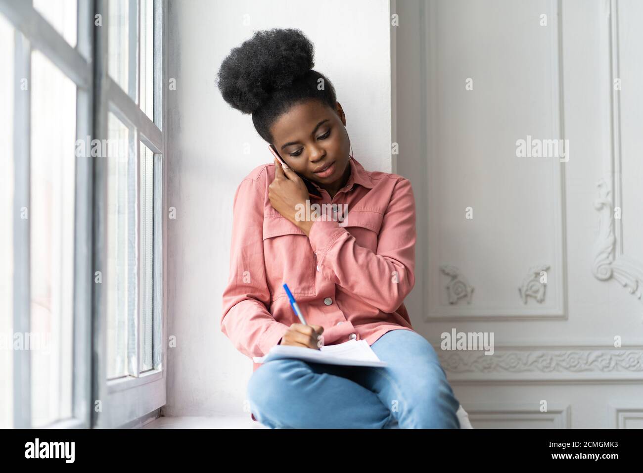 Fokussierte afroamerikanische Biracial Frau Notizen machen, klärt Informationen, Telefonieren, sitzen auf Fensterbank. Biracial weibliche Mitarbeiter konsultieren Stockfoto