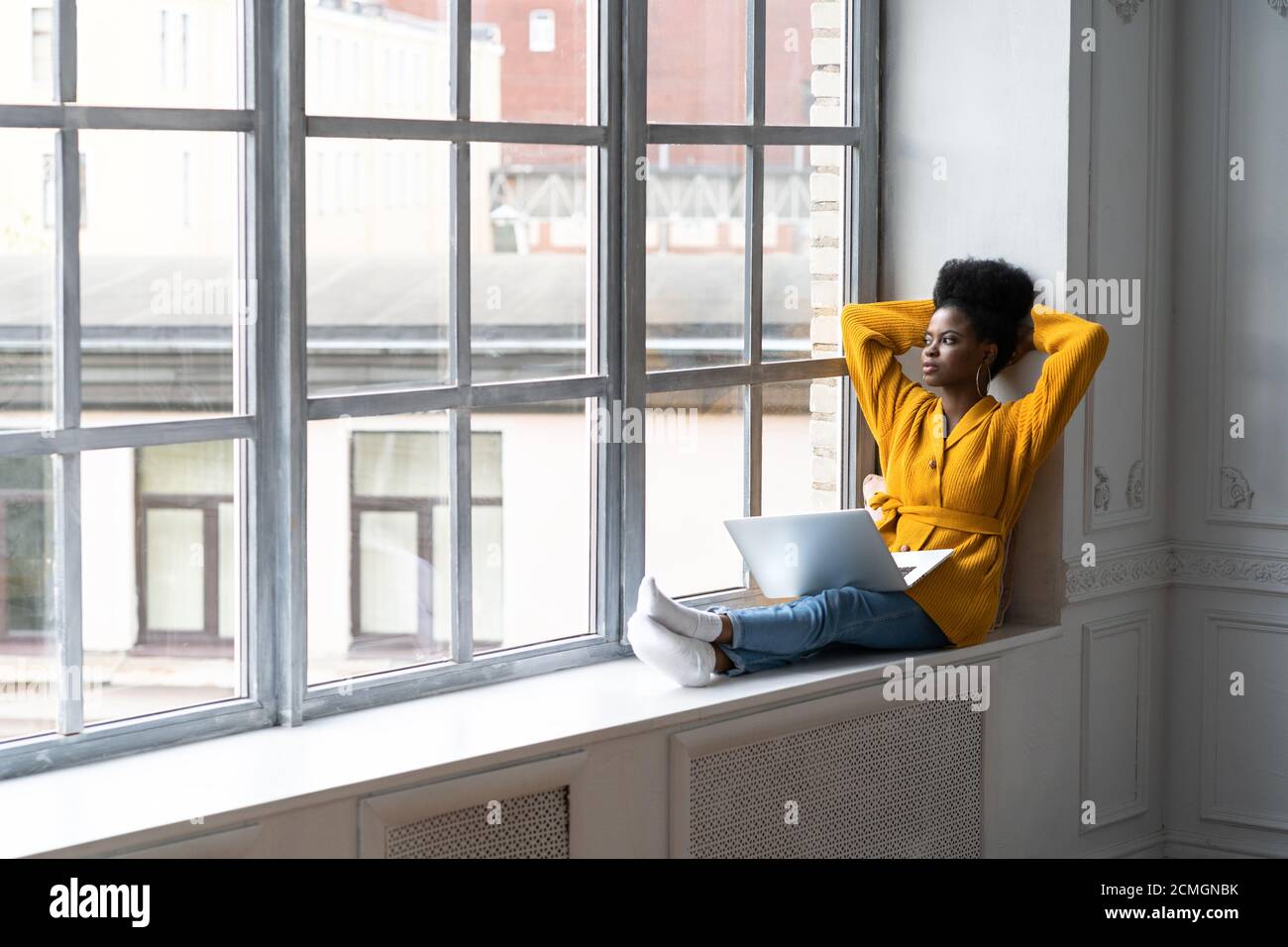 Entspannte afroamerikanische Millennial Frau mit Afro-Frisur tragen gelbe Strickjacke, sitzen auf Fensterbank, ruhen, Pause von der Arbeit auf Laptop, Stockfoto