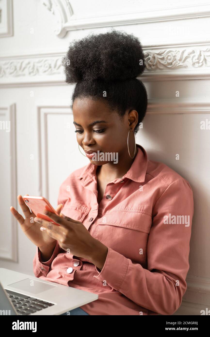 Nahaufnahme Porträt der afroamerikanischen tausendjährigen jungen Frau in trendigen Schmuck große Ohrringe tragen rosa Hemd sitzen, mit Smartphone und Eingabe messa Stockfoto