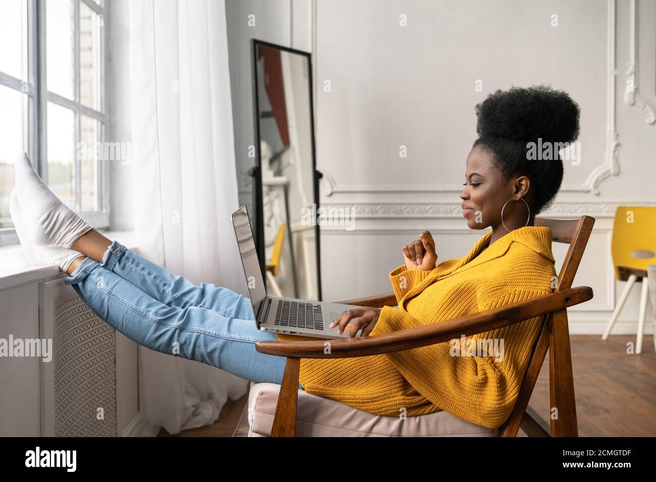 Happy African American Frau Freiberuflerin mit afro Frisur tragen gelbe Strickjacke ruhen, sitzen auf Stuhl, Beine auf Fensterbank lehnend, Remote-Studyin Stockfoto