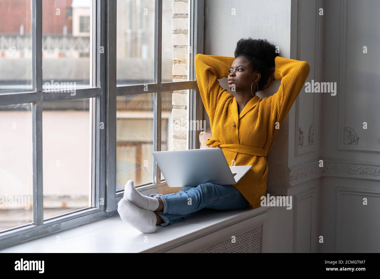Entspannte afroamerikanische Millennial Frau mit Afro-Frisur tragen gelbe Strickjacke, sitzen auf Fensterbank, ruhen, Pause von der Arbeit auf Laptop, Stockfoto