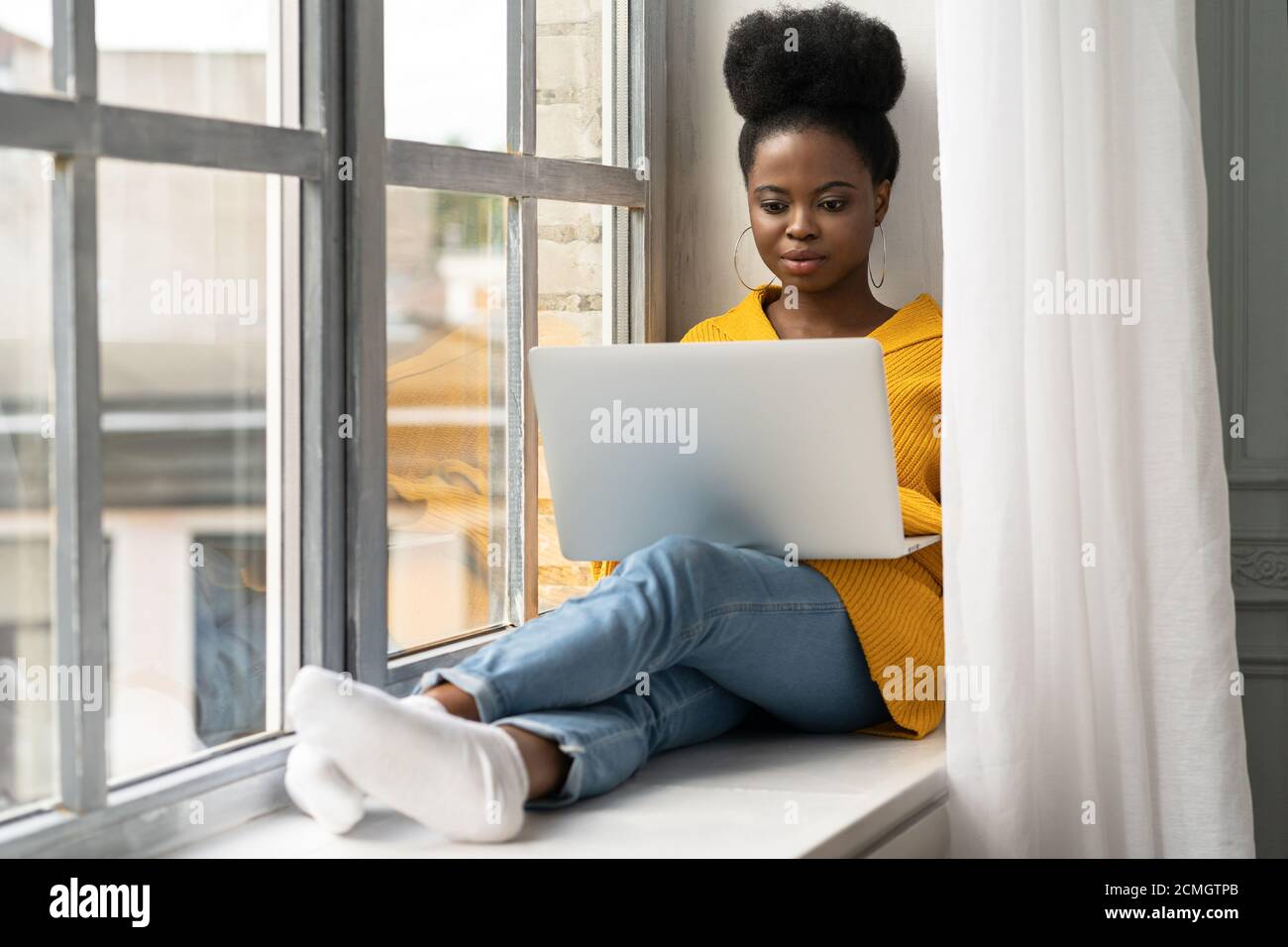 Afroamerikanische Studentin mit Afro-Frisur tragen gelbe Strickjacke, sitzen auf Fensterbank, arbeiten Remote-Job auf Laptop, Lernen mit onl Stockfoto