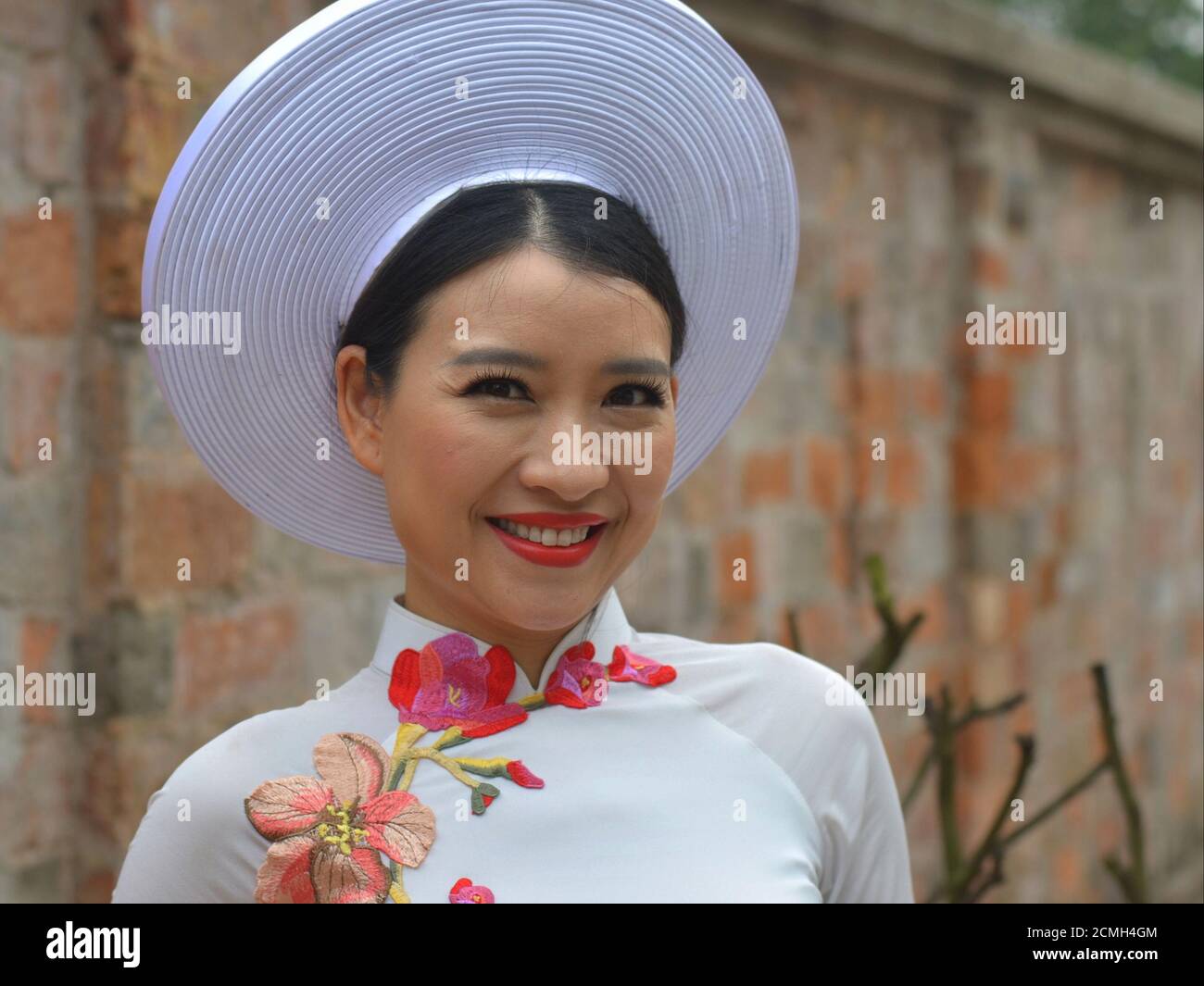 Schöne vietnamesische Frau trägt ein besticktes weißes Brautkleid und einen traditionellen vietnamesischen Brauthut und lächelt für die Kamera. Stockfoto