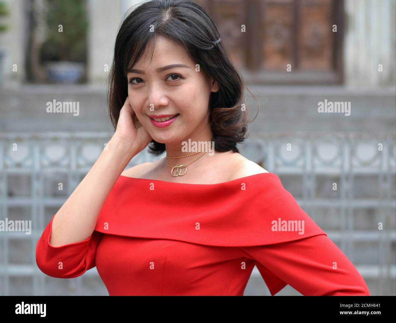 Schöne junge lächelnde vietnamesische Geschäftsfrau mit schulterlangen braunen Haaren trägt ein selbst maßgeschneidertes rotes Kleid und schaut auf die Kamera. Stockfoto