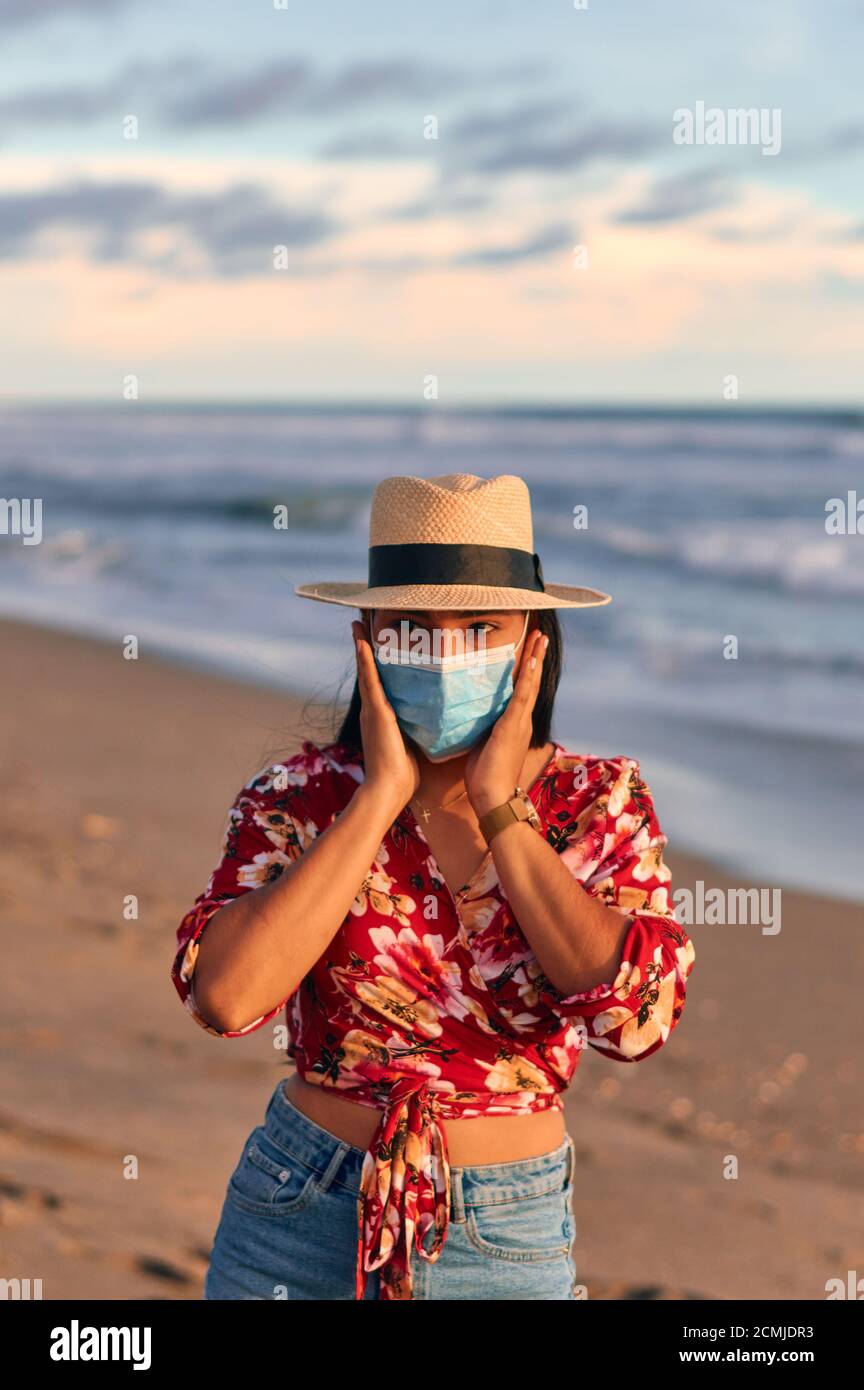 Eine junge Frau, die ein schönes rotes Hemd mit Blumenmustern, Shorts und Hut trägt, steht auf der Küste und trägt eine Gesichtsmaske, um covid-19. Hol zu verhindern Stockfoto