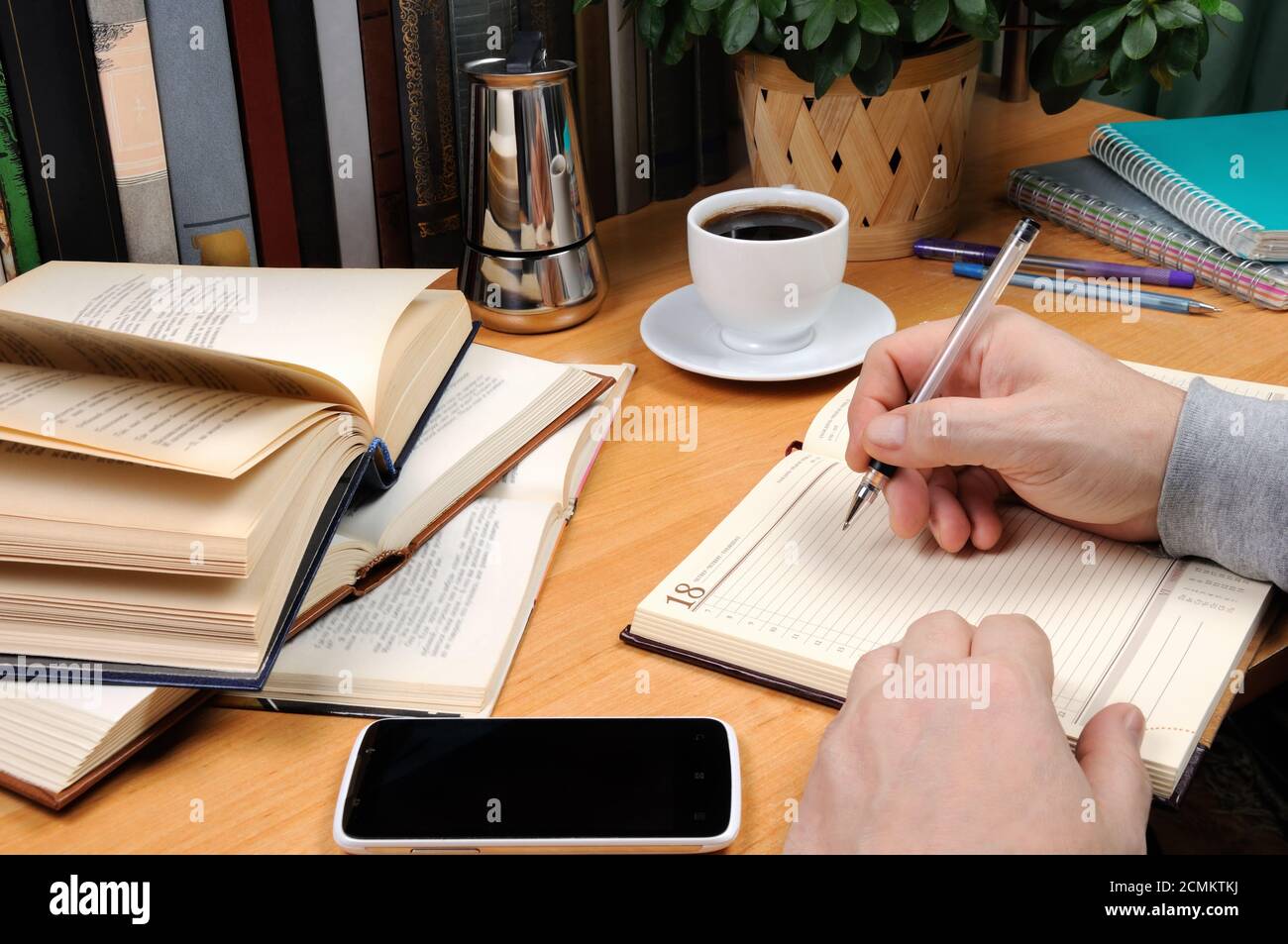 Ein Mann hält einen Stift und Notizen in einem Notizbuch auf einem Schreibtisch in seinem Büro, umgeben von Büchern, notebo Stockfoto