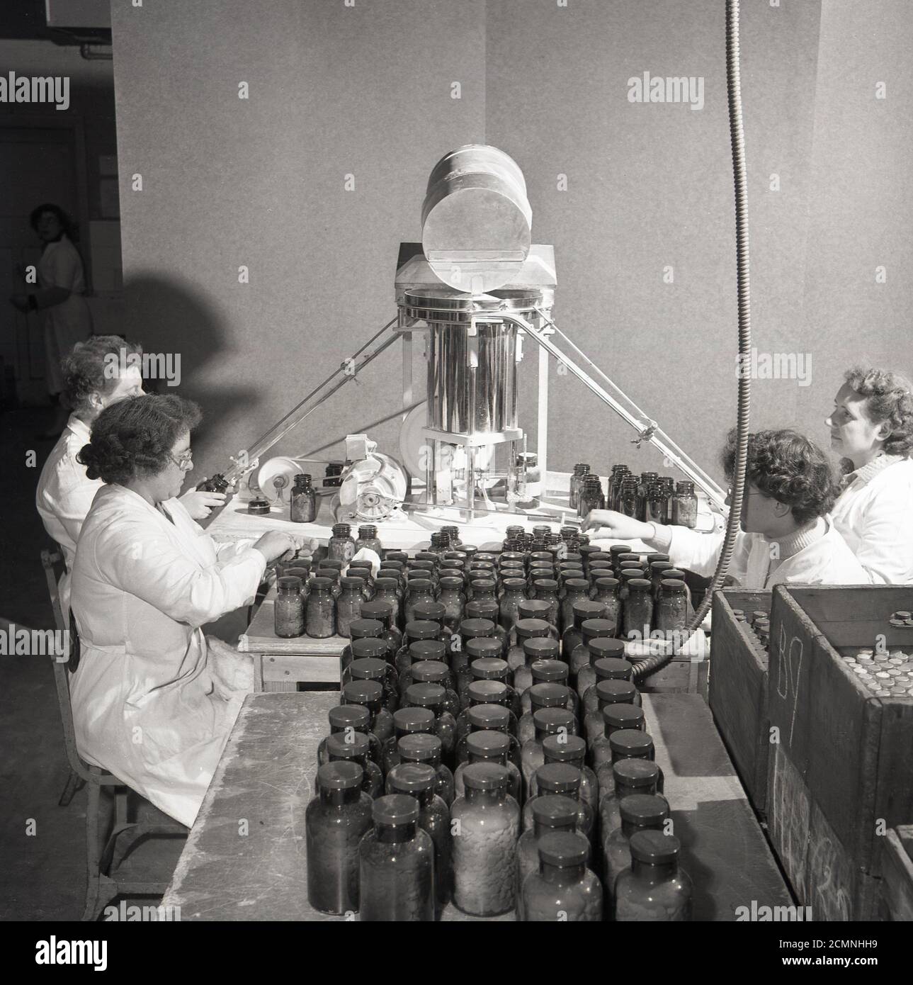 1950er Jahre, historisches Bild von J Allan Cash von vier weiß gestrichenen Arbeiterinnen bei der medizinischen oder pharmatualen Produktionsfirma Arthur Cox & Sons. Zwei der Damen sitzen mit Maschinen, um die Glasflaschen zu versiegeln, und neben ihnen setzen zwei Damen Plastikdeckel auf die Flaschen. Stockfoto