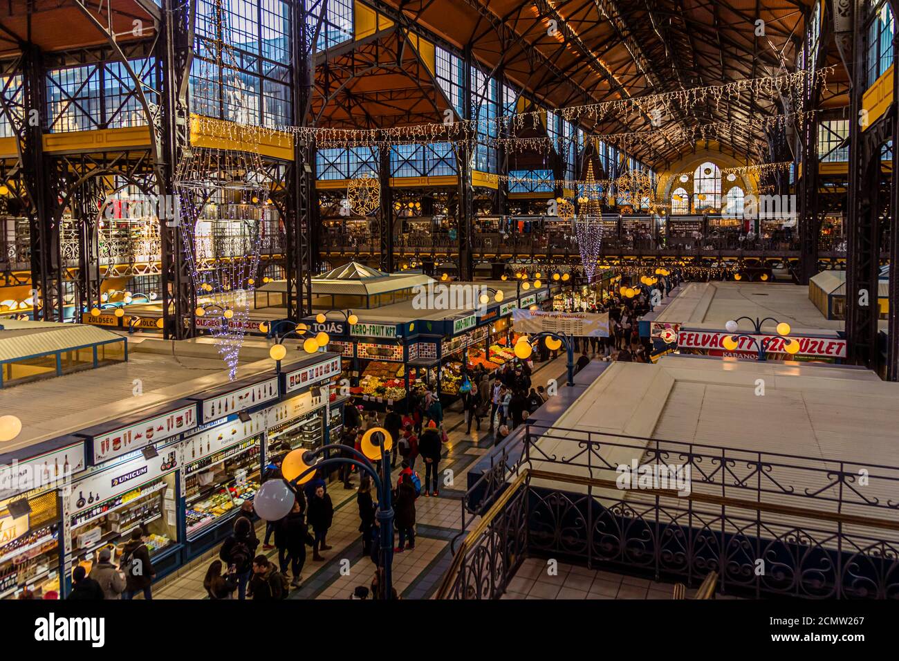 Lichterketten und Weihnachtskugeln hängen zwischen den grünen Stahlträgern der Großen Markthalle im 9. Bezirk von Budapest, Ungarn, während Besucher durch die Gänge zwischen den Ständen schlendern. Der Architekt Samu Pecz baute die Halle, die 1897 eröffnet und mit glasierten Zsolnay-Kacheln verkleidet wurde; sie war bis 1932 die größte Markthalle der Stadt. Banner mit historischen Fotos hängen von der oberen Galerie. Stockfoto