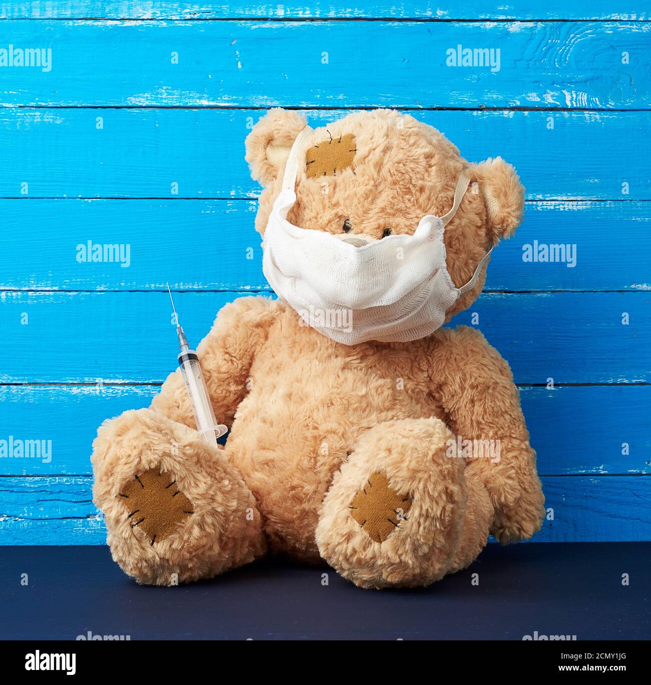 Beige große Teddybär mit Patches sitzen in einem weißen Medizinische Maske in der Pfote einer Spritze Stockfoto