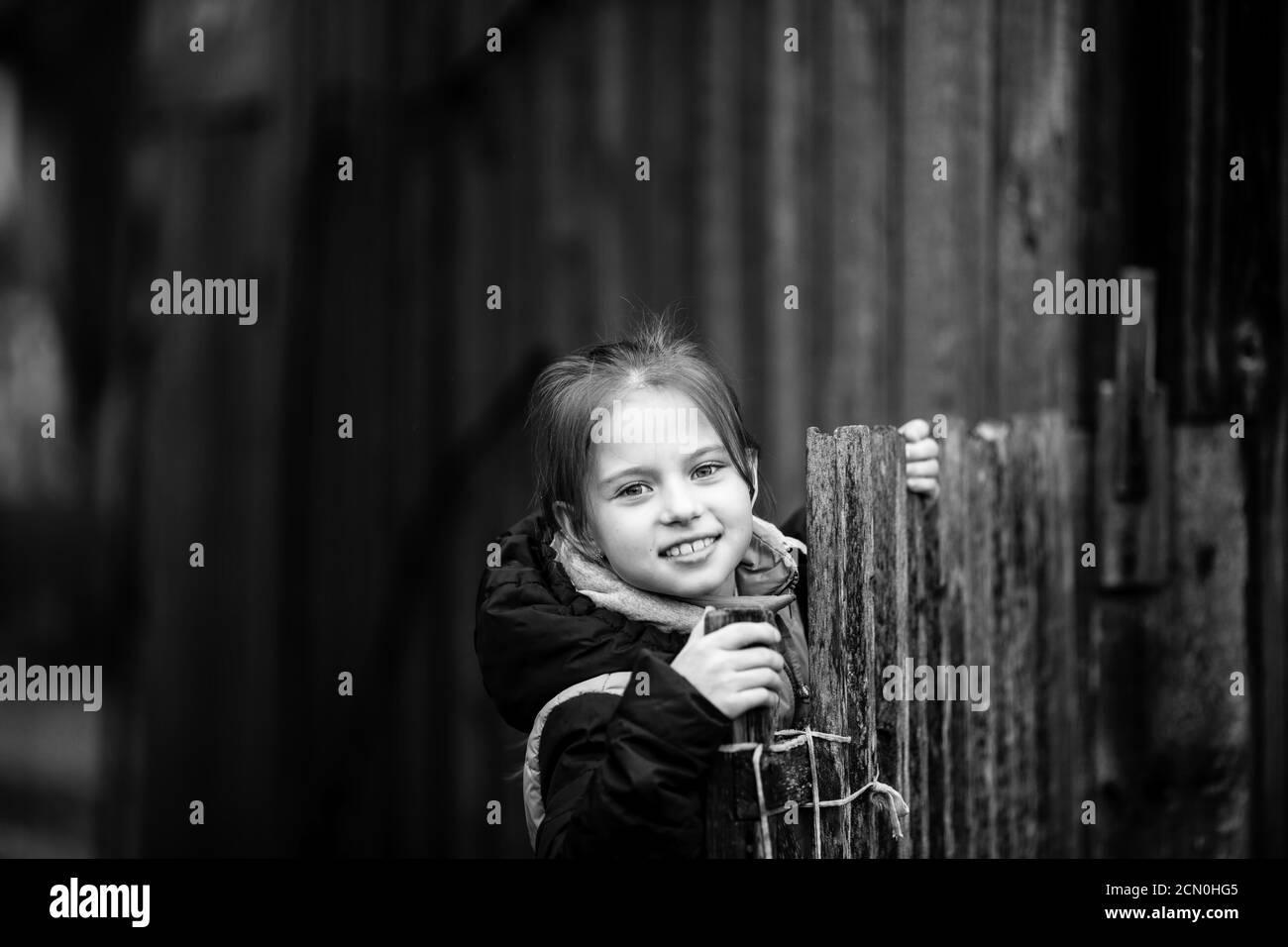 Porträt eines kleinen Mädchens hinter einem Holzzaun in einem Dorf. Schwarzweiß-Fotografie. Stockfoto