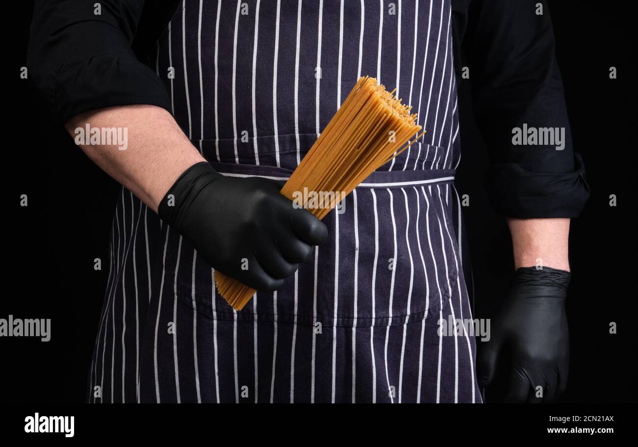 Chefkoch in schwarzen Latexhandschuhen, gestreifte Schürze hält roh lange gelbe Spaghetti aus Hartweizen Stockfoto