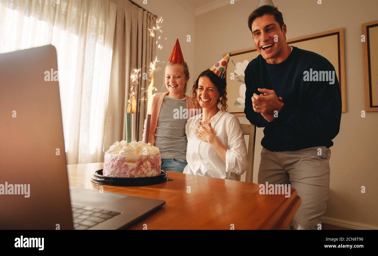 Frau feiert Geburtstag zu Hause mit Familie und Freunden auf Videoanruf. Laptop und Geburtstagskuchen mit Kerzen auf dem Tisch mit Familie trägt Party Hut Stockfoto