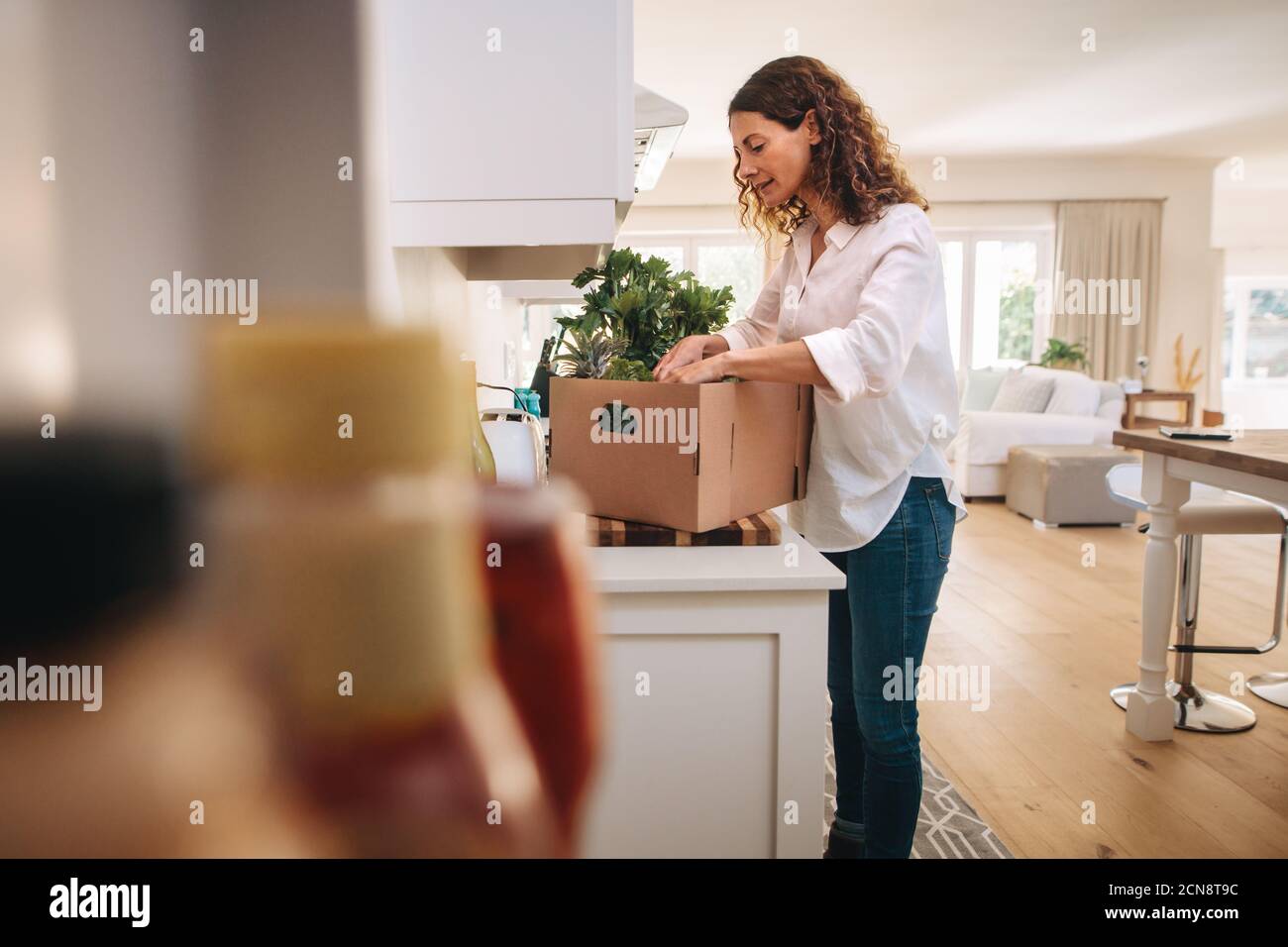 Frau, die in der Schachtel mit Lebensmitteln von der Lieferung in ihrer Küche. Frau steht in der Küche und überprüft die Früchte und Gemüse in der Lieferbox. Stockfoto