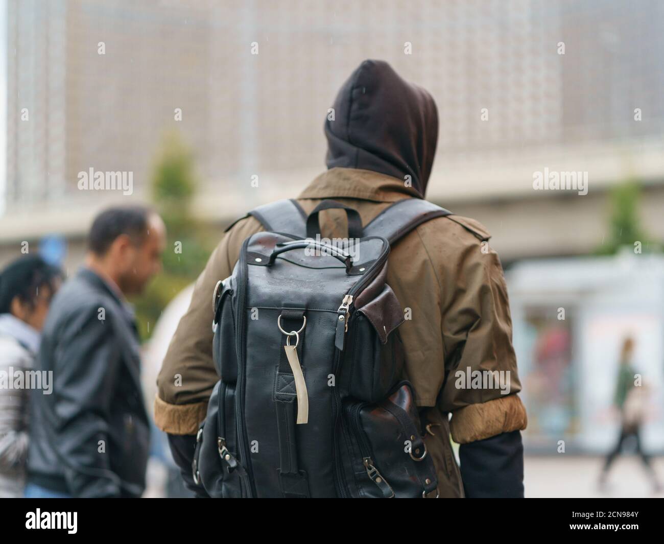 Junger Mann mit schwarzem Rucksack an der herbstlichen Stadtstraße. Regentag. Er zog Kapuzenpullover und Regenmantel an. Moderne Mode Stil der Jugend. Rückansicht/Rückansicht. C Stockfoto