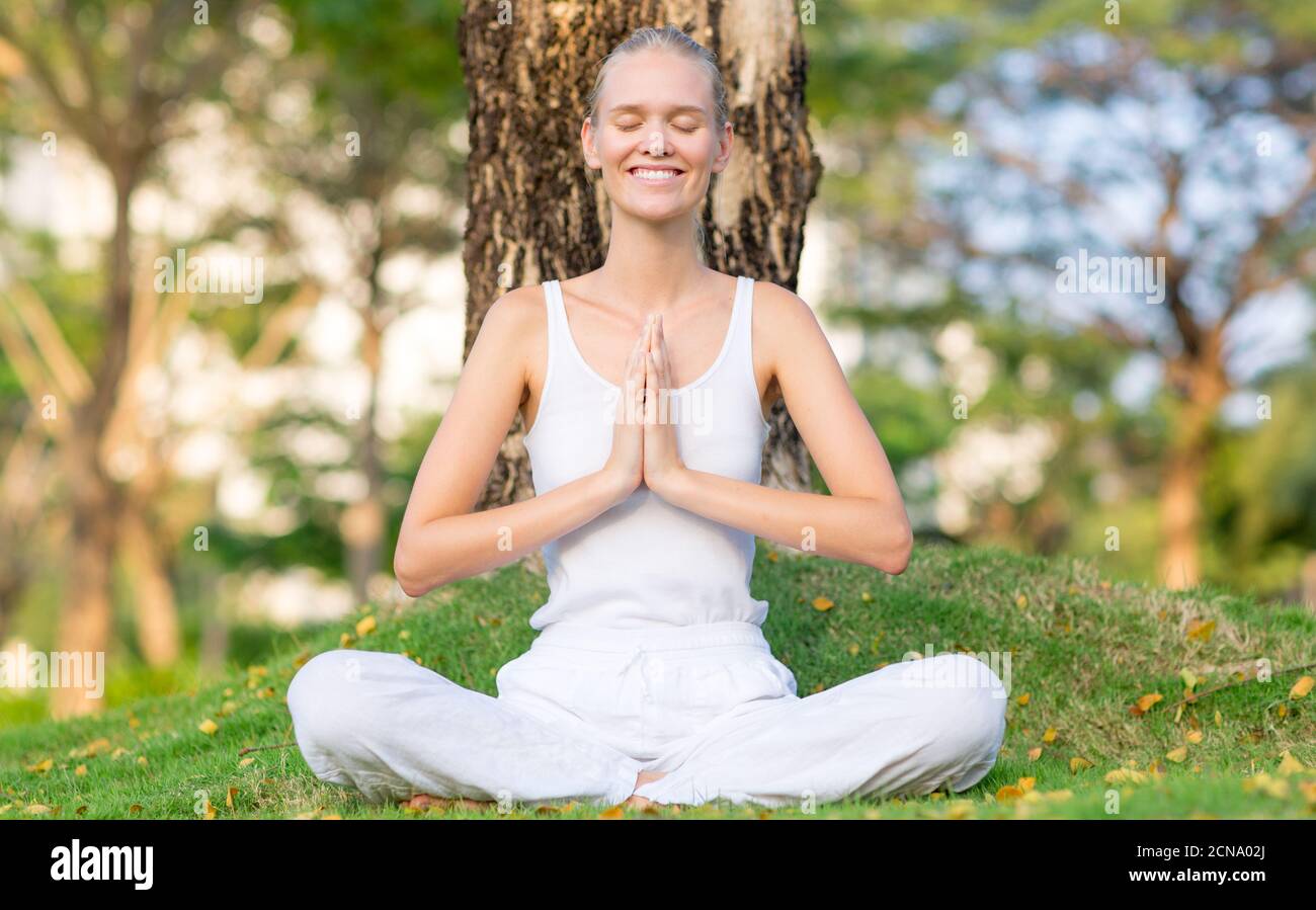 Wohlbefinden, Gesundheit und Selbstversorgung. Glückliche Frau, die Yoga praktiziert und meditiert, sitzt auf dem Gras in der Natur mit Bäumen im Hintergrund. Stockfoto