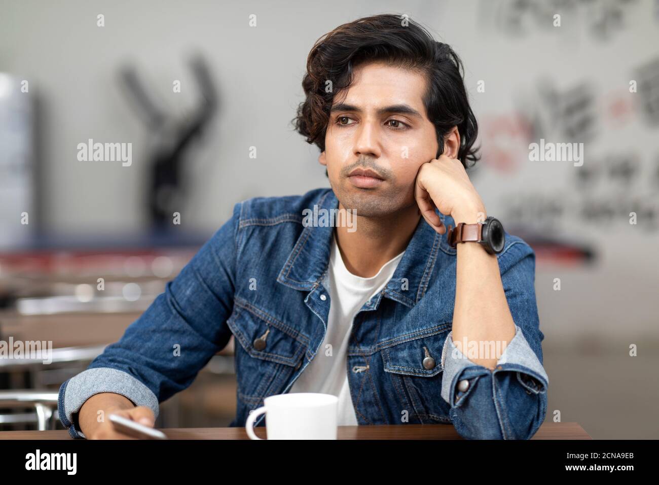Porträt eines jungen, gutaussehenden indischen Mannes mit ernstem Blick, verloren in Gedanken, sitzt in einer Bürokantine, Café, lässige Arbeitsumgebung. Stockfoto