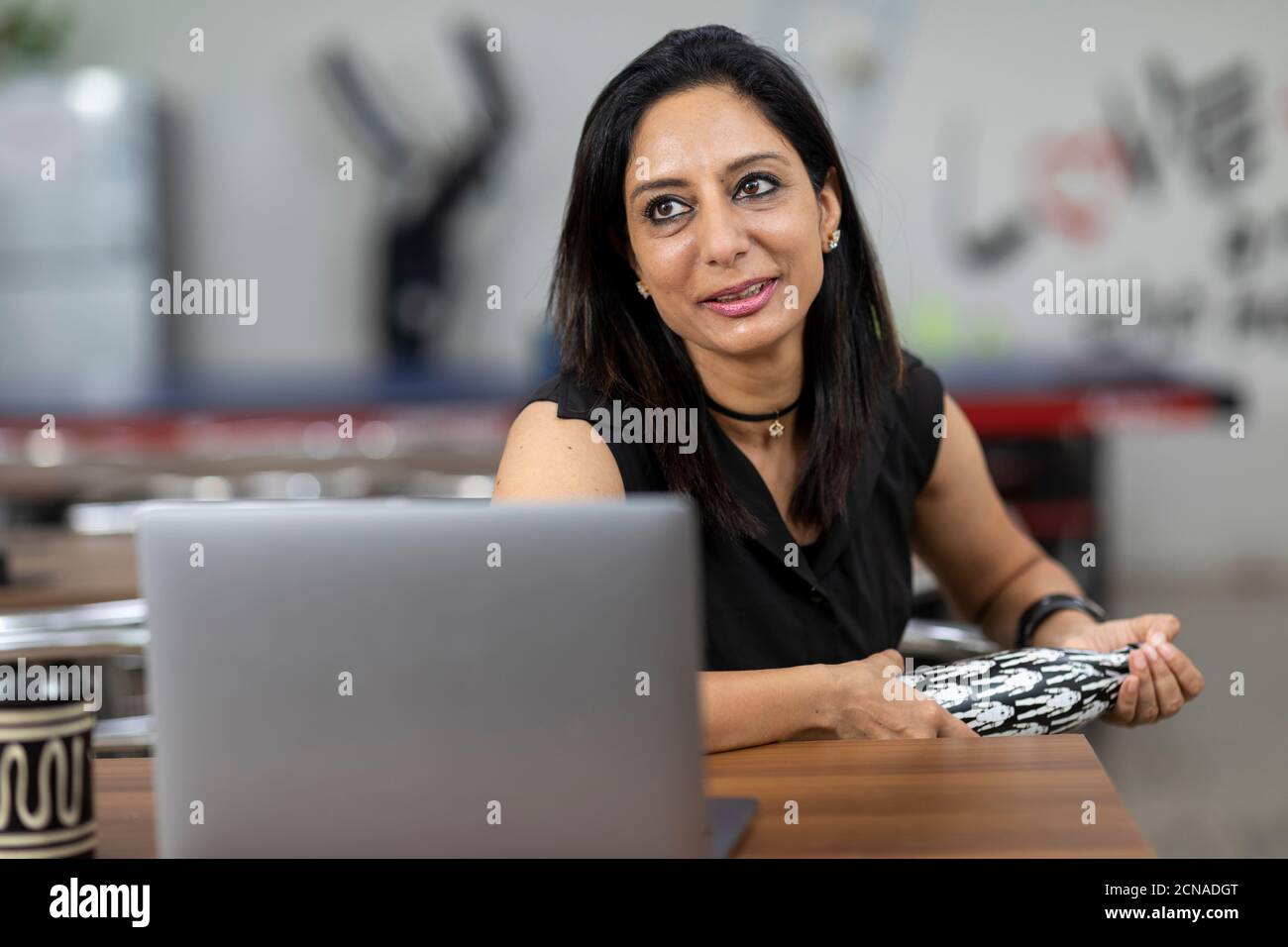 Porträt einer lächelnden indischen Frau vor ihrem Laptop, sitzt in einer Bürokantine, Café, zwanglose Arbeitsumgebung. Stockfoto