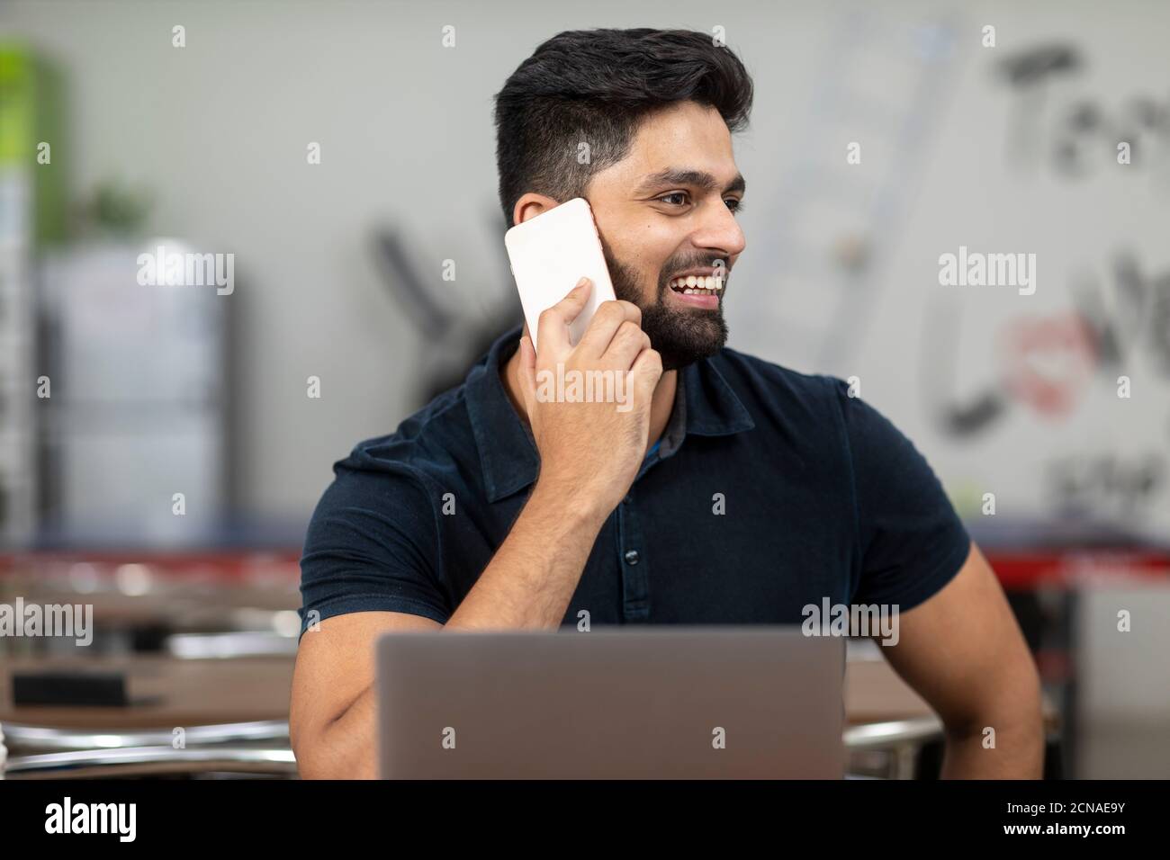 Porträt eines jungen hübschen indischen Mannes, der am Telefon spricht und am Laptop arbeitet, in einer Bürokantine sitzt, in einem Café, in einer legeren Arbeit. Stockfoto
