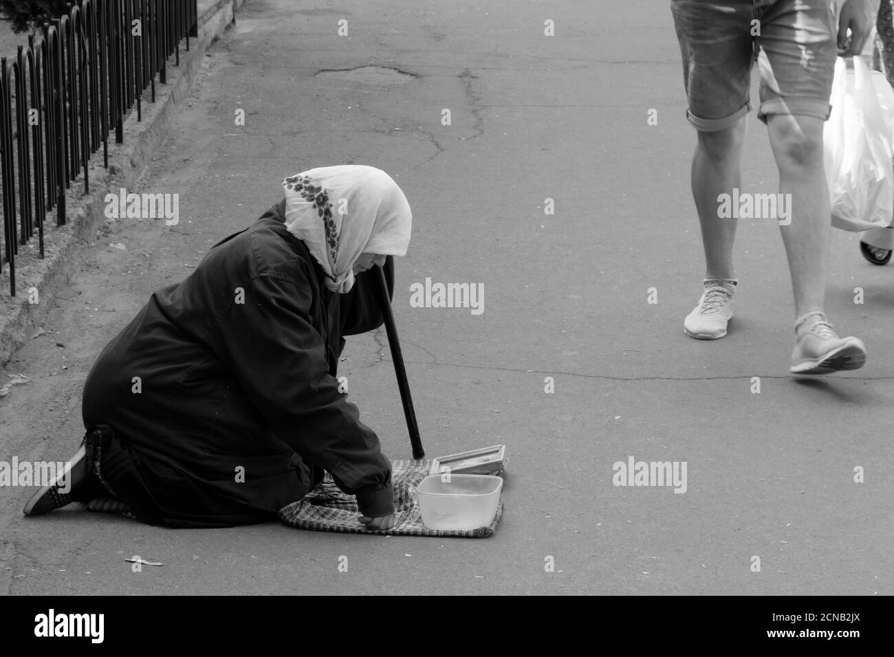 Tschernihiw, Ukraine, 7. Juni 2019. Eine ältere Frau in einem Schal betteln um Geld und kniet auf einer Stadtstraße. Ein Typ in Shorts geht an einem Bettler vorbei. Monochrom Stockfoto