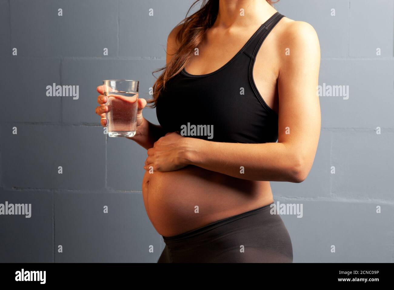 Schwangere Frau im Sport-Workout-Outfit mit einem Glas Wasser. Eine Nahaufnahme des Bauches einer schwangeren Frau. Gesunde Ernährung während der Schwangerschaft. Stockfoto