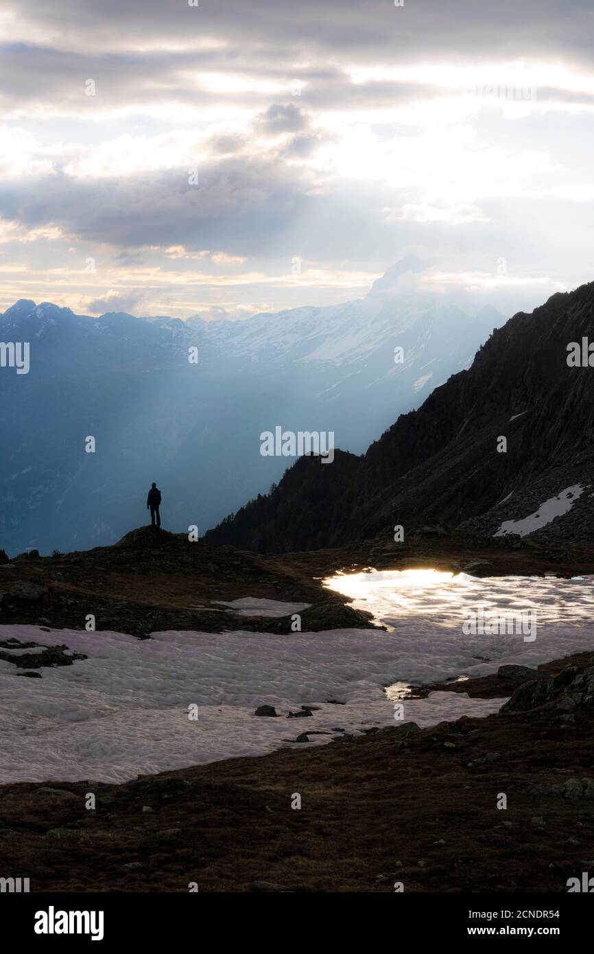 Silhouette eines Menschen, der bei Sonnenaufgang am Ufer des Sees Zana, Valmalenco, Valtellina, Lombardei, Italien, Europa unter Sonnenstrahlen steht Stockfoto