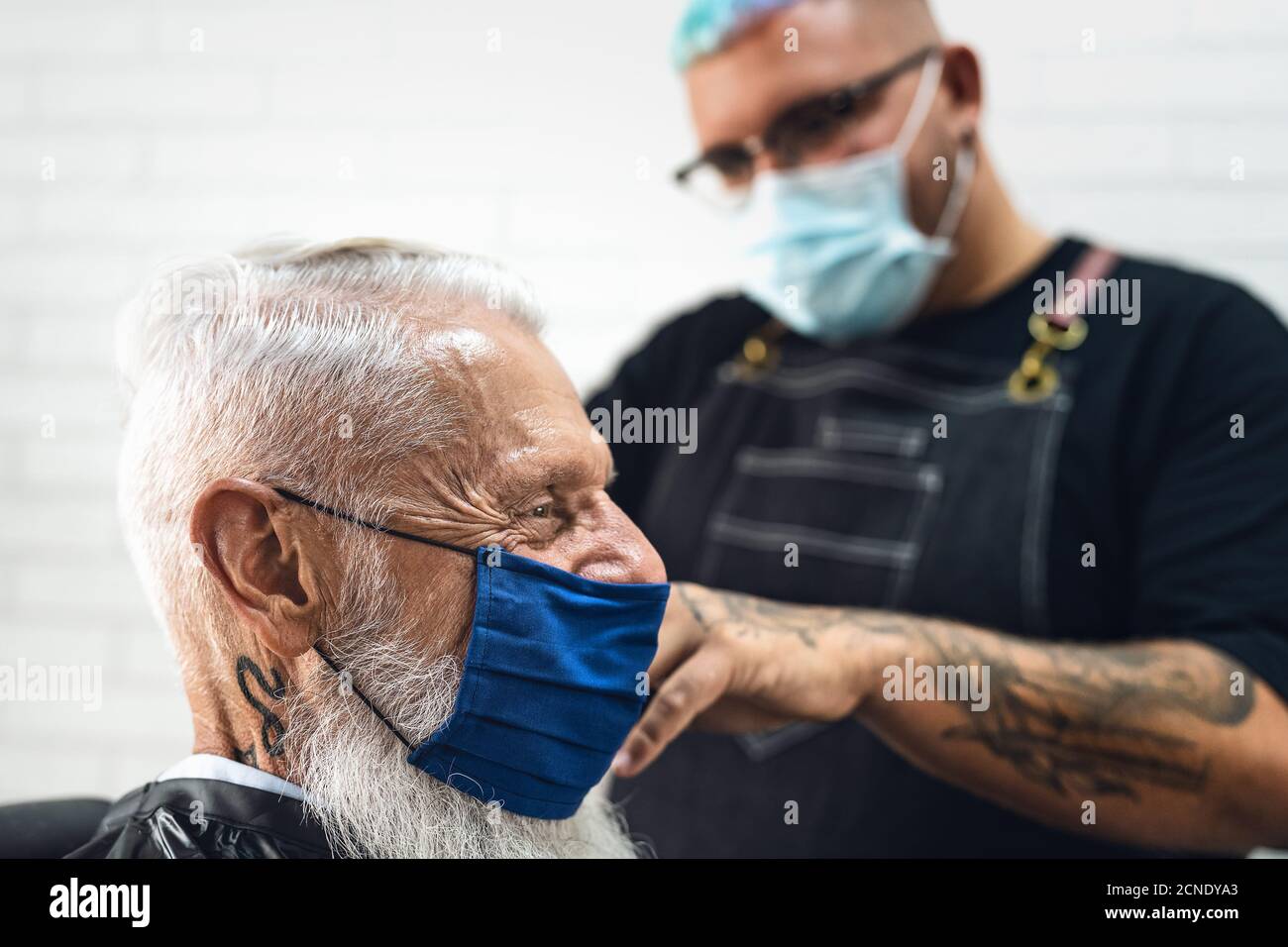 Männliche Friseur schneiden Haare zu Hipster Senior Client während des Tragens Gesicht chirurgische Maske - Junge Friseurin arbeitet in Friseursalon während corona-Virus Stockfoto