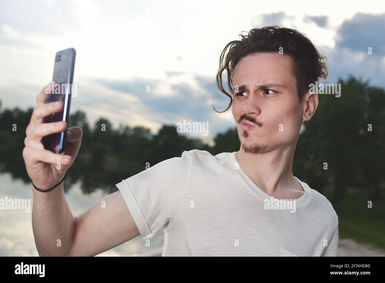 Frustrierter junger gutaussehender Kerl macht erfolgloses Selfie auf seinem Smartphone in der Natur, der Kerl Grimassen in die Kamera.der Mann re Stockfoto