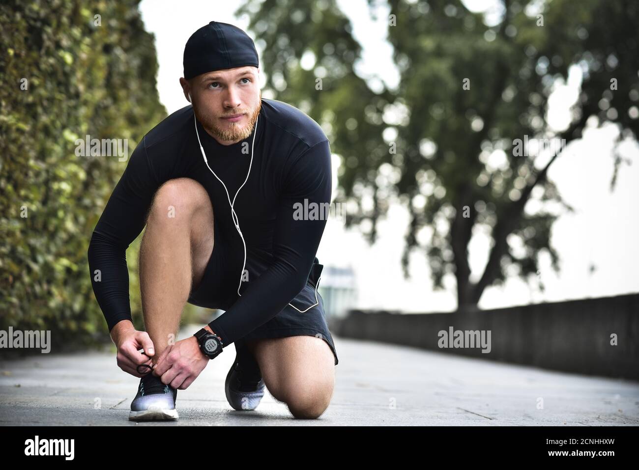 Ernsthafter bärtiger kaukasischer junger Sportler, der im Stadtpark Schnürsenkel auf seine Sneakers bindet. Stockfoto