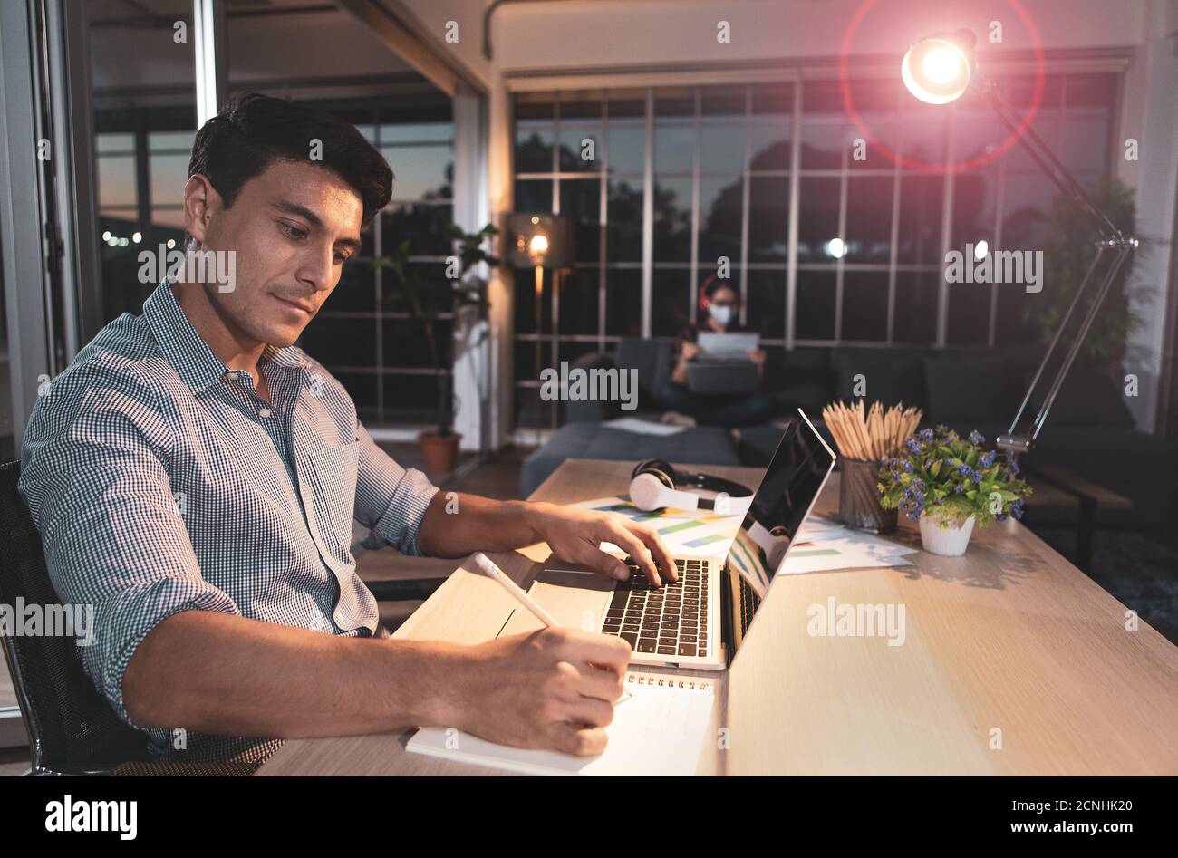 Kaukasischer Geschäftsmann sitzt glücklich auf dem Schreibtisch im Wohnzimmer unter warmen Lampe mit Fackel cicle und Dämmerung blauen Himmel außerhalb des Hauses. Idee für die Arbeit la Stockfoto