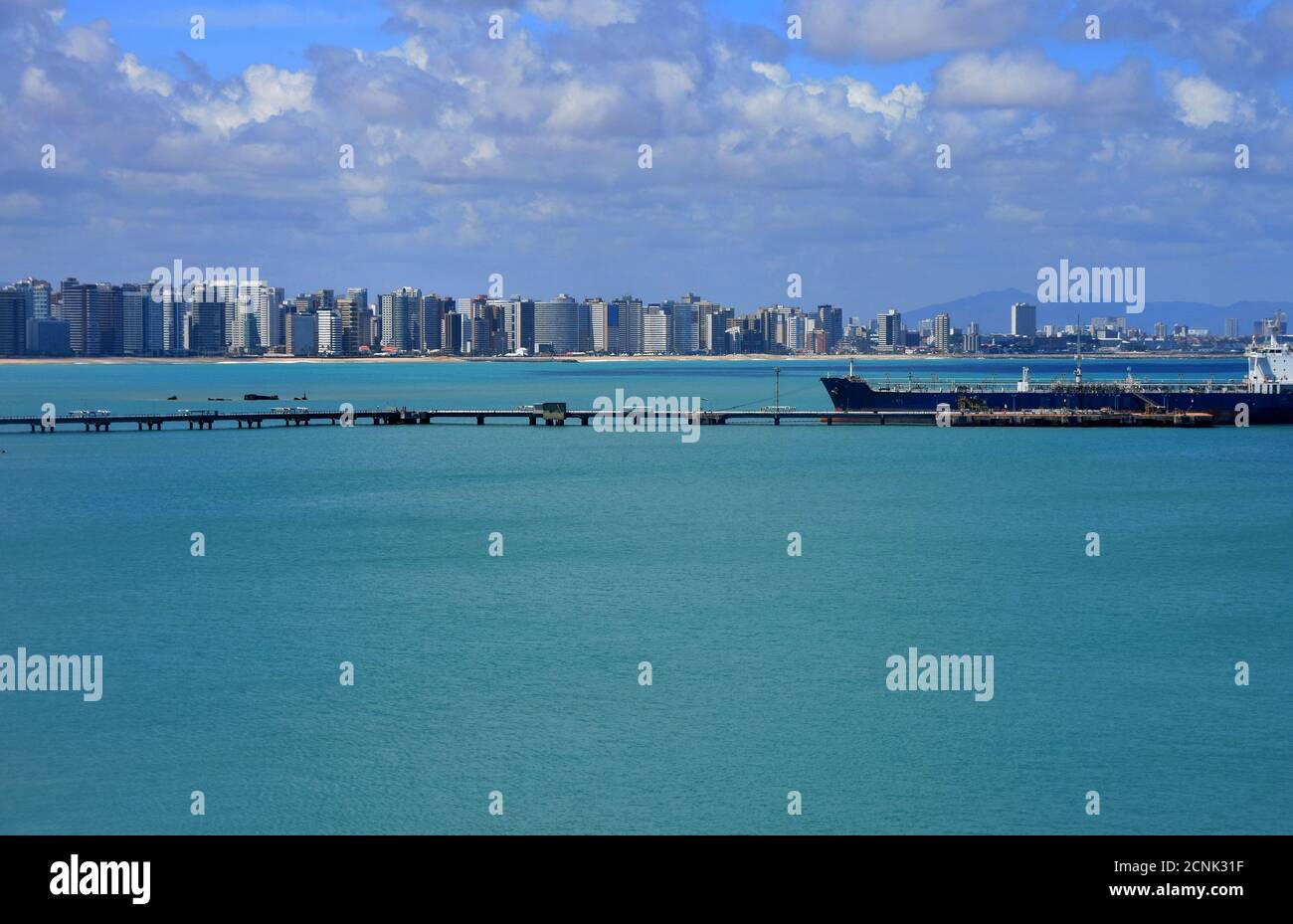 Stadtbild von Port Fortaleza in Brasilien während des Sommertages unter azurblauem Himmel mit Wolken und ruhiger Oberfläche des Atlantiks. Im Vordergrund blauer Tankwagen. Stockfoto
