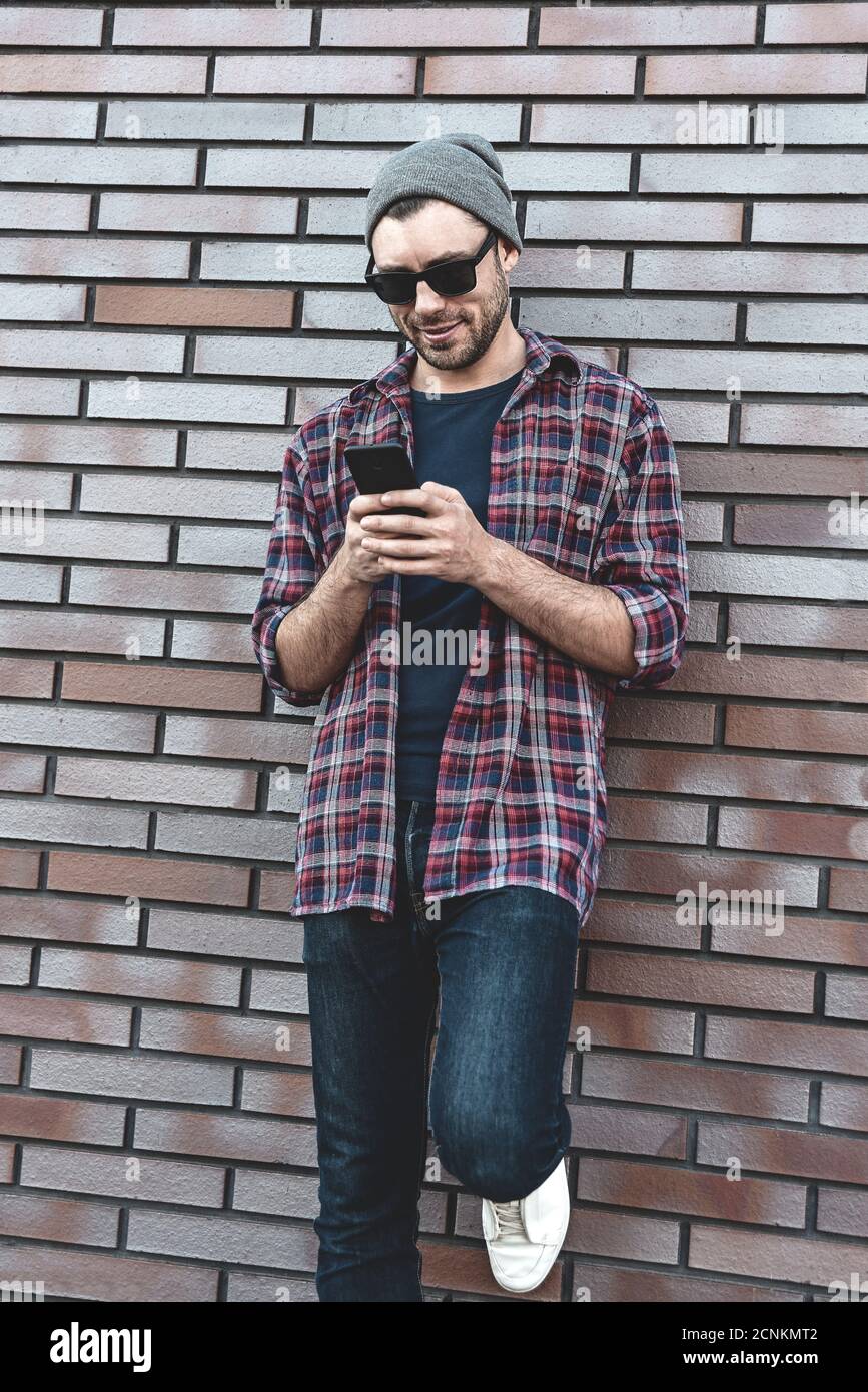 Textnachricht eingeben. Seitenansicht des hübschen jungen Mannes in eleganter Freizeitkleidung, der Mobiltelefon hält, während er sich an der Ziegelwand lehnt. Stockfoto