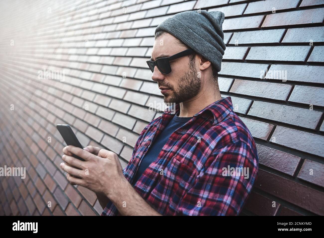 Textnachricht eingeben. Seitenansicht des hübschen jungen Mannes in eleganter Freizeitkleidung, der Mobiltelefon hält, während er sich an der Ziegelwand lehnt. Stockfoto