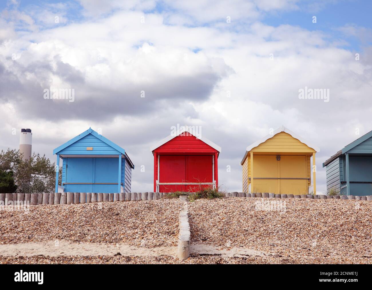 Primäre farbige, rote, gelbe, blaue Reihe von Strandhütten und Kraftwerk Schornstein, am englischen Strand am Meer, Calshot, Hampshire. Stockfoto