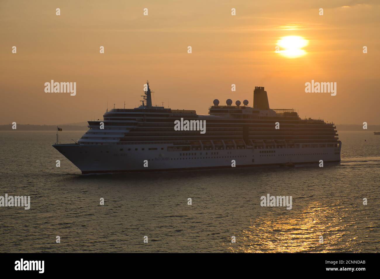 Ein Kreuzfahrtschiff, das bei Sonnenuntergang in den Hafen von Portsmouth segelt, wobei die untergehende Sonne einen Lichtweg auf der Wasseroberfläche mit dem Liner in Silhouette macht Stockfoto