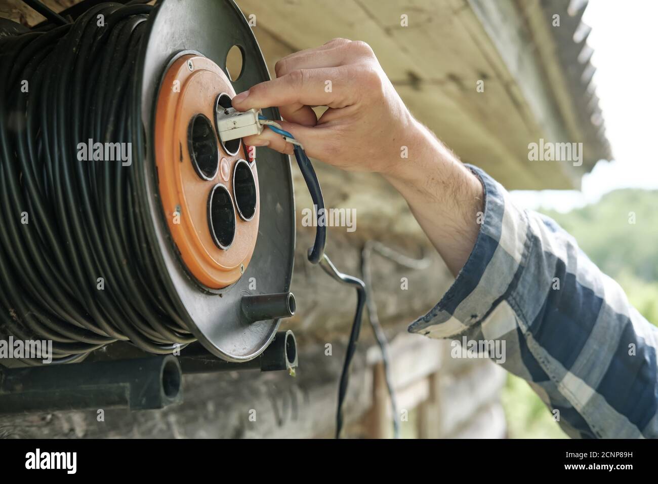 Eine männliche Hand steckt einen Stecker in eine Steckdose, die unter dem Dach einer alten Scheune in einem ländlichen Innenhof hängt. Nahaufnahme. Stockfoto