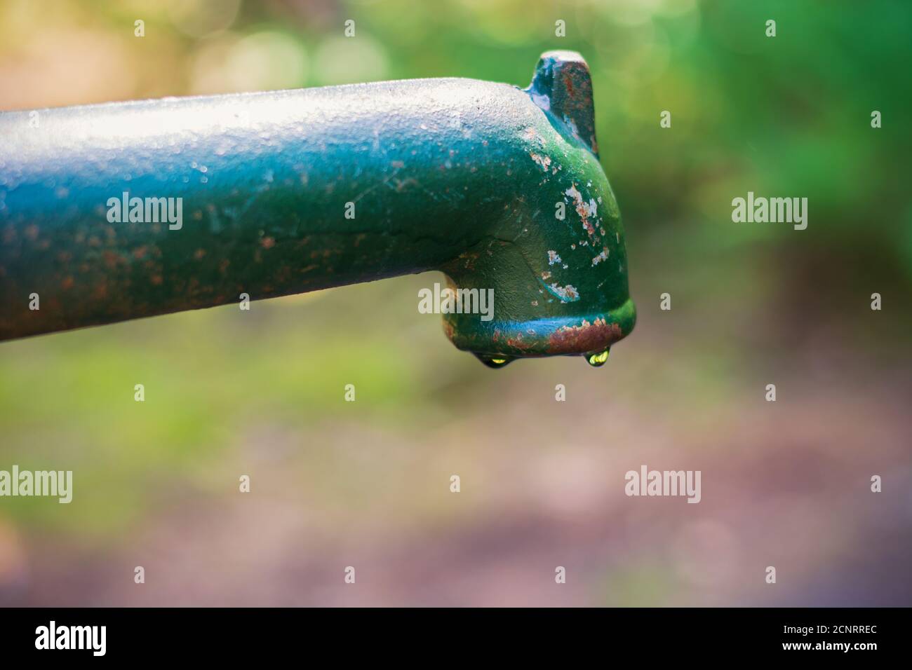 Manuelle alte grüne und rostige Wasserhebelpumpe mit Wasser aus dem Auslauf, letzten Tropfen Wasser, Trockenheit. Verschwommener grüner Hintergrund Stockfoto