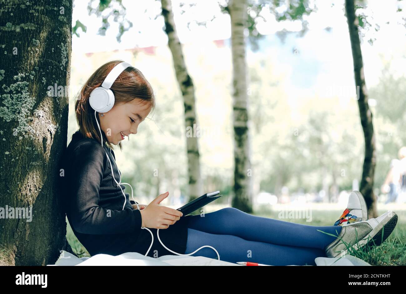 Glückliches Mädchen in weißen Kopfhörern mit digitalen Tablet-pc im Park. Fernlernkonzept. Resilienz, zurück zur Schule, neue Normalität. Stockfoto