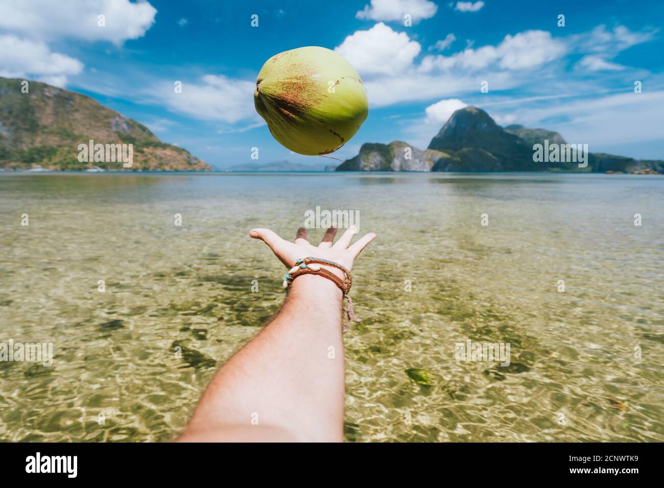 POV jonglieren mit Kokosnuss in männlichen Händen gegen exotische Ozeanlagune und Insel im Hintergrund. Stockfoto