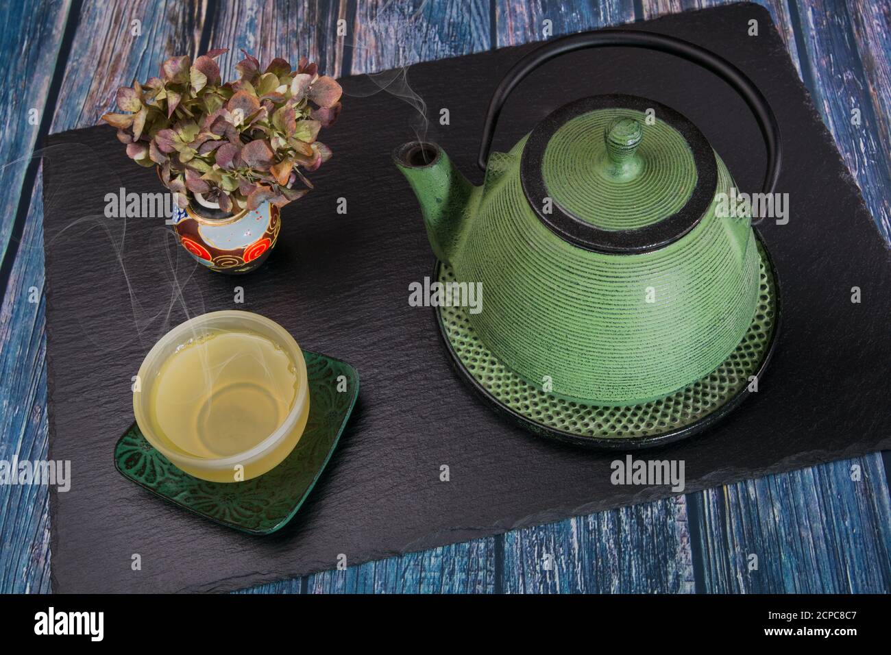 Eine dampfende Teekanne aus grünem Metall, ein Glasaufsatz mit dampfendem Tee und eine chinesische Vase mit getrockneten Hortensien auf einer Schieferplatte. Stockfoto