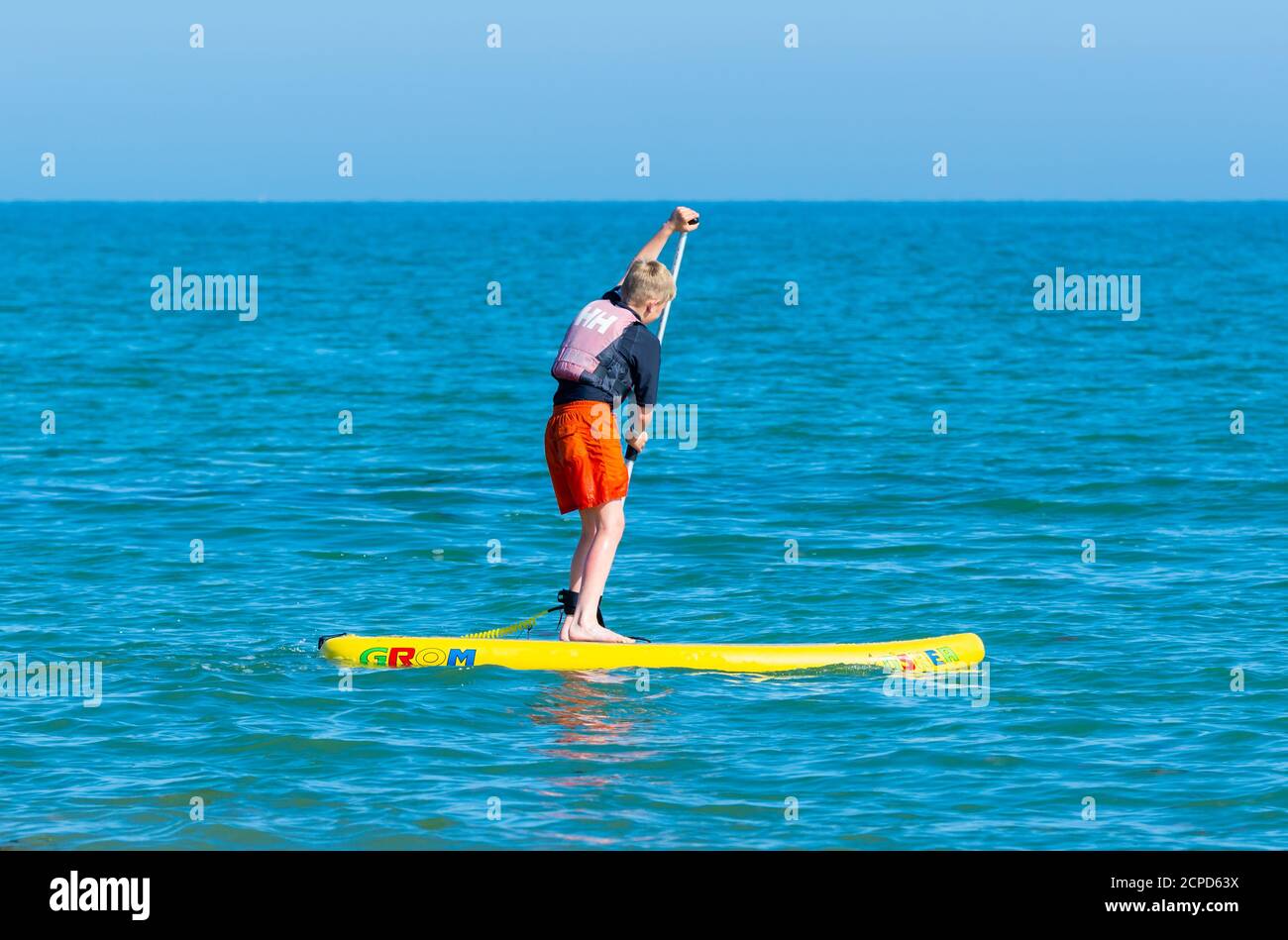 Ein Junge, der auf einem Paddelbrett steht, paddelt an einem heißen Sommertag in England, Großbritannien, auf See. Stockfoto