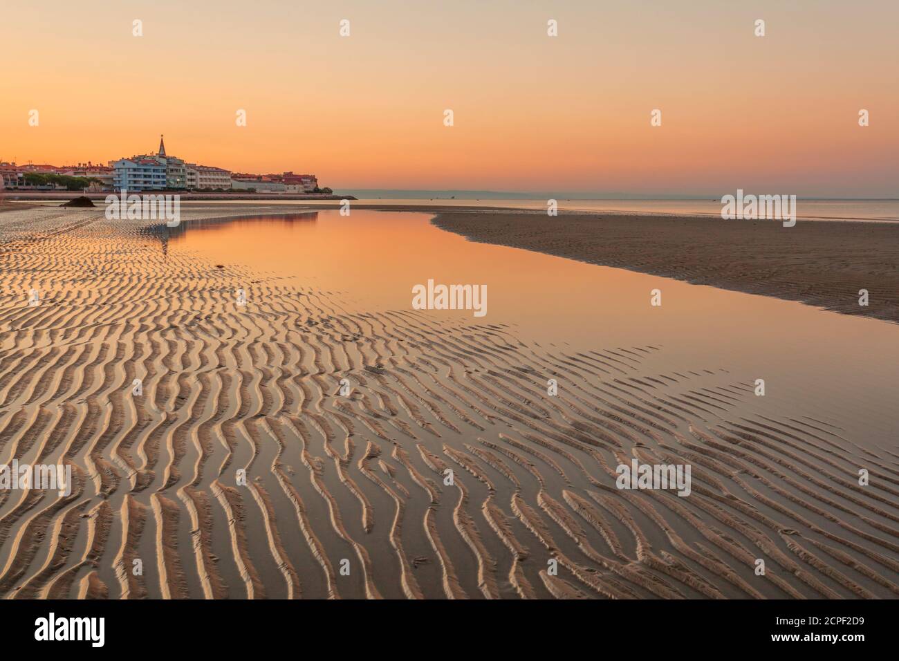 Farbenfroher orangefarbener Strand bei Sonnenuntergang mit Wellensandmustern aus Die rückläufigen Gezeiten und Reflexionen auf dem Wasser der Lagune in einer ruhigen Landschaft Stockfoto