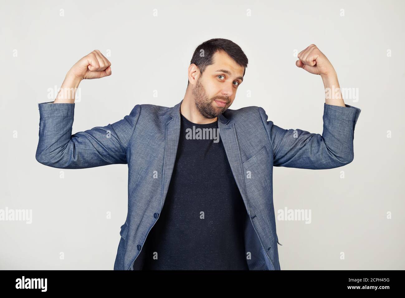 Junger lächelnder Geschäftsmann mit Bart in der Jacke, der die Armmuskeln zeigt und stolz lächelt. Fitnesskonzept. Porträt eines Mannes auf grauem Hintergrund. Stockfoto