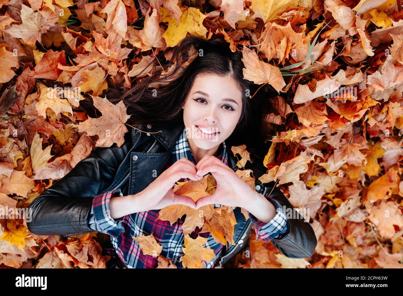 Ich liebe den Herbst. Junge lächelnde Frau, die auf Herbstblättern liegt und Herz macht Stockfoto