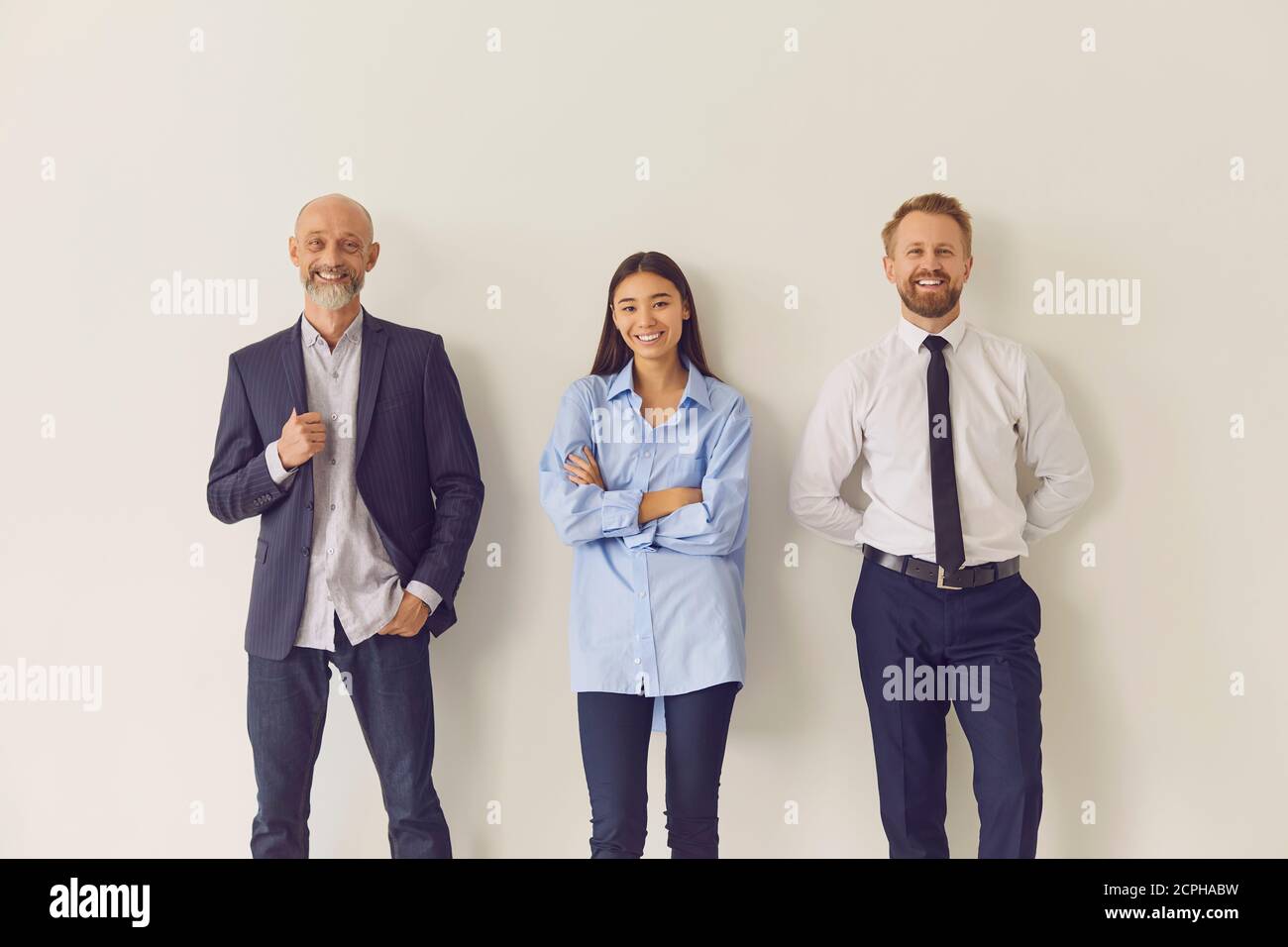 Drei fröhliche, mehrjährige Büroangestellte, die sich an die Wand lehnen und schauen Bei der Kamera Stockfoto