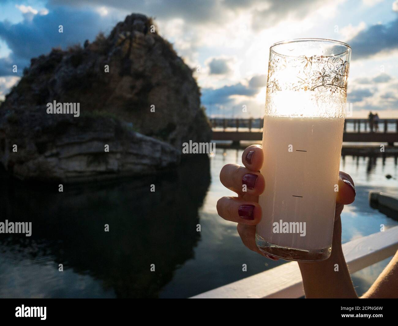 Eine nagelpolierte weibliche Hand hält ein Glas Getränk für einen Jubel in einem Restaurant am Wasser. Ein großer Felsen spiegelt sich am Meer und die Sonne ist Stockfoto